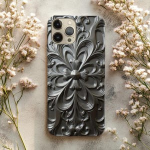 May include: A grey phone case with an elaborate, floral pattern. The case has a detailed, raised design of swirling leaves and a central flower-like motif. The case has a glossy finish and a camera cutout.