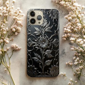 May include: Black phone case with a detailed floral design in silver. The case features raised flowers, leaves, and ornate scrollwork. The phone's camera lenses are visible at the top. The case is surrounded by delicate white flowers.