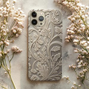 May include: A light gray phone case with a detailed, raised floral pattern. The case features a camera cutout and is surrounded by delicate white flowers, creating a soft, elegant aesthetic. The design includes leaves and swirling vines.