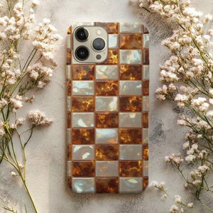Può includere: Una custodia per telefono con un motivo a scacchi di quadrati marroni e bianchi. La custodia ha una finitura lucida e un ritaglio per la fotocamera nella parte superiore. Il design ricorda il guscio di tartaruga. Fiori bianchi sullo sfondo.