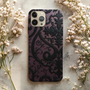 May include: A mobile phone case featuring a dark purple backdrop and a black, elaborate floral pattern. The case has a raised texture and a camera aperture. White flowers surround the case.