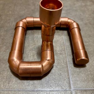 May include: A copper pipe candle holder with a single candle holder on top. The holder is made of copper pipe fittings and is in the shape of a U.