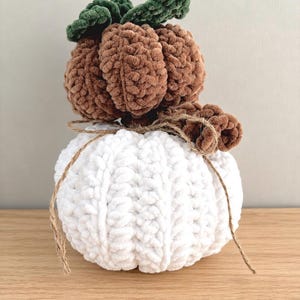 May include: A stacked pumpkin decoration made of soft, chunky yarn. The bottom pumpkin is white, and the top pumpkin is brown. A dark green crocheted leaf sits on top, and a thin brown twine is tied around the middle, featuring a small cinnamon stick.