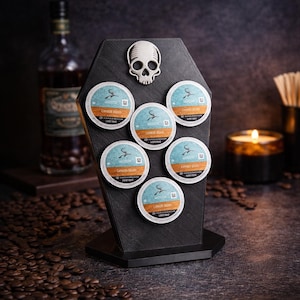 May include: A white ceramic mug with a black handle and the text "But First, Coffee" in a bold, black font. The mug is set against a blurred background, suggesting a cozy setting.