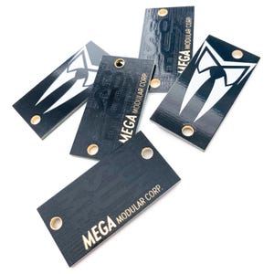 May include: A collection of black rectangular tags with gold eyelets. Some tags feature a white tuxedo design, while others display the text "MEGA MODULAR CORP." in gold. The tags are scattered against a white background.
