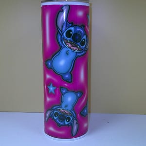 Puede incluir: Un vaso alto y rosa con un personaje de dibujos animados azul. El personaje se muestra en dos poses, una vertical y otra invertida, con una estrella azul. El vaso tiene un borde y una base blancos.