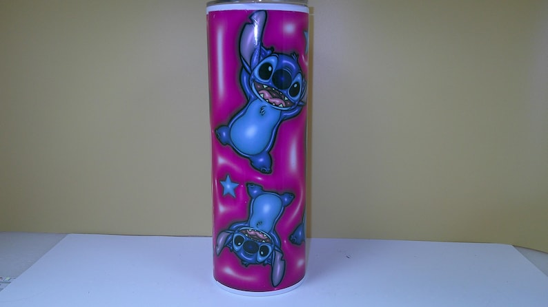 Puede incluir: Un vaso alto y rosa con un personaje de dibujos animados azul. El personaje se muestra en dos poses, una vertical y otra invertida, con una estrella azul. El vaso tiene un borde y una base blancos.
