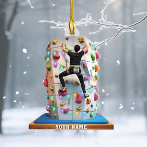 May include: A decorative ornament featuring a rock climbing wall with a person climbing. The wall is covered in colorful climbing holds and lights. The base has the text "YOUR NAME" on a wooden platform. The ornament hangs from a gold string.