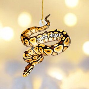 May include: A decorative ornament shaped like a ball python snake, featuring a detailed pattern of brown, black, and yellow markings. The text "YOUR NAME" is printed on the snake's body. The ornament is suspended by a gold string.
