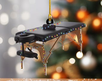May include: A black massage table ornament with a headrest, armrests, and a rolled towel. The ornament is decorated with colorful string lights and a gold hanging cord. The words "Your Name" are written on the side.