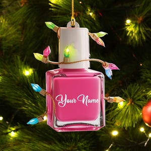 May include: A pink nail polish bottle-shaped Christmas ornament with the text "Your Name" in white script. The ornament is decorated with a string of colorful Christmas lights and hangs from a gold string.