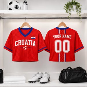 a pair of red Croatia soccer jerseys hanging on a rack, with the word "Croatia" printed on the front. The jerseys also have the text "Your Name" and the number "00" printed on the back. In addition to the jerseys, there are various sports equipment items such as a soccer ball, a water bottle, and a pair of sneakers.
