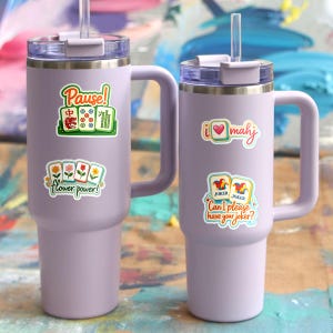 Two purple insulated tumblers with colorful stickers on them, placed on a colorful background.