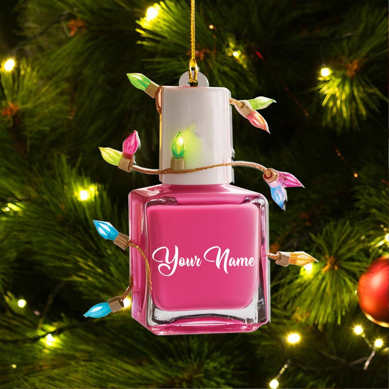 May include: A pink nail polish bottle-shaped Christmas ornament with the text "Your Name" in white script. The ornament is decorated with a string of colorful Christmas lights and hangs from a gold string.