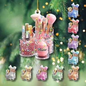 May include: A clear acrylic makeup brush holder with multiple compartments, filled with pink beads, makeup brushes, and lipsticks. The holder has the year "2024" and the name "Name" printed on it. Additional smaller makeup brush holders are also shown.