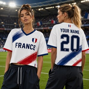 Two women wearing white and red soccer jerseys with the word "France" on the front and the number "20" on the back, standing on a soccer field.