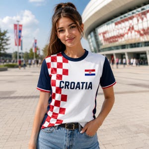 A young woman wearing a white and red Croatia soccer jersey stands in front of a stadium, smiling at the camera.