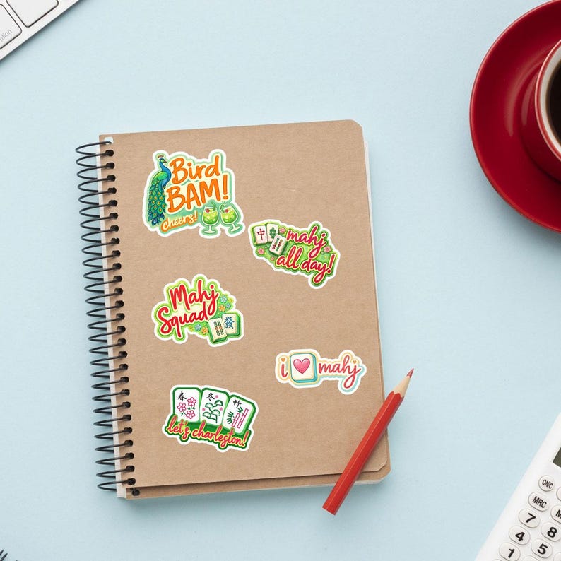 a spiral-bound notebook with various stickers on the cover, a red cup, a red pen, and a calculator on a light blue background.
