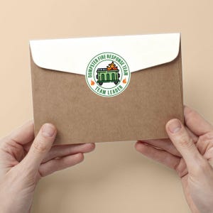May include: A brown envelope with a white flap is held by two hands. A circular sticker on the envelope reads "Dumpster Fire Response Team" around a green dumpster with flames and the words "Team Leader".