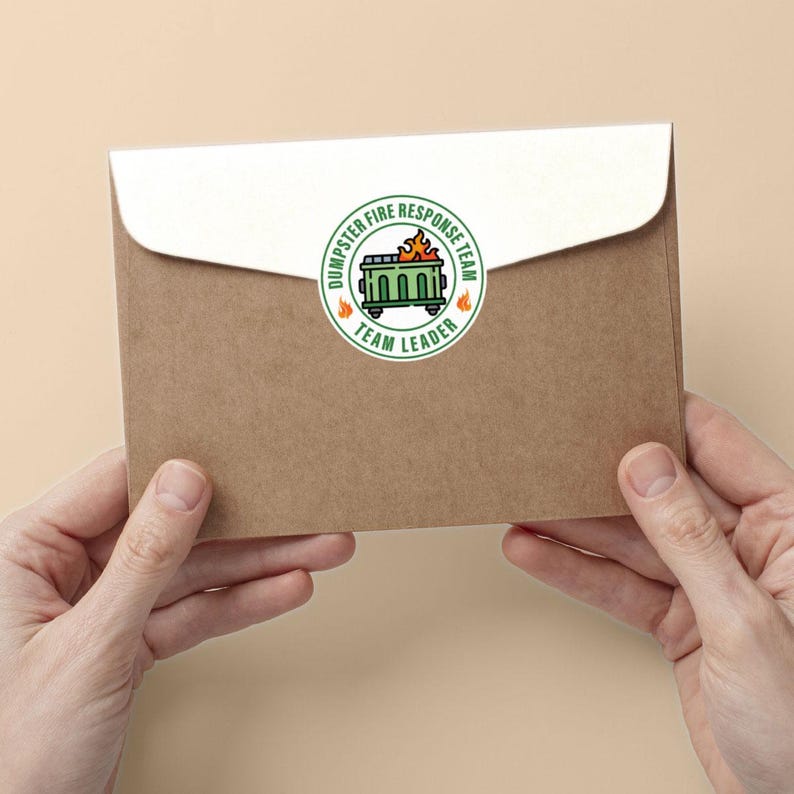 May include: A brown envelope with a white flap is held by two hands. A circular sticker on the envelope reads "Dumpster Fire Response Team" around a green dumpster with flames and the words "Team Leader".