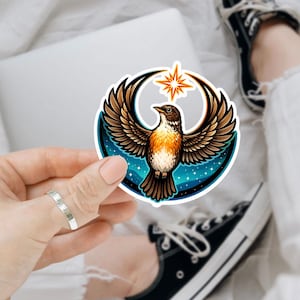 May include: A sticker featuring a stylized bird with outstretched wings, set against a crescent moon and starburst. The bird has brown and orange feathers, with a blue and starry background. The sticker is held by a hand.