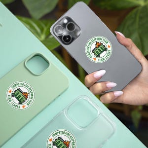 May include: A collection of phone cases and a phone, each featuring a circular sticker. The sticker design includes a green dumpster on fire, with the text "DUMPSTER FIRE RESPONSE TEAM" and "TEAM LEADER" in a circular layout. The phone cases are in green and clear.