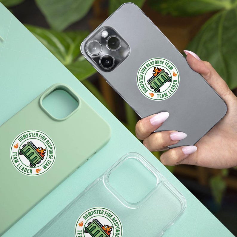 May include: A collection of phone cases and a phone, each featuring a circular sticker. The sticker design includes a green dumpster on fire, with the text "DUMPSTER FIRE RESPONSE TEAM" and "TEAM LEADER" in a circular layout. The phone cases are in green and clear.