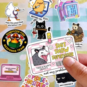 a person holding a sticker with a cartoon image of a raccoon and the text "SHIT SHOW SUPERVISOR" along with a humorous message about being tired and always on duty.