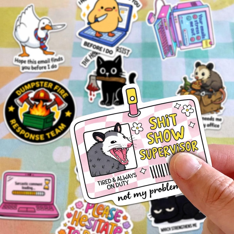 a person holding a sticker with a cartoon image of a raccoon and the text "SHIT SHOW SUPERVISOR" along with a humorous message about being tired and always on duty.