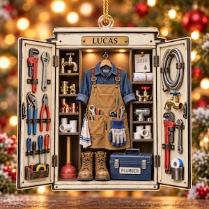 May include: A decorative ornament shaped like an open cabinet labeled LUCAS, featuring a miniature denim shirt, brown apron, work boots, and a blue toolbox labeled PLUMBER. The interior displays various plumbing tools, brass fittings, and a 2026 calendar.