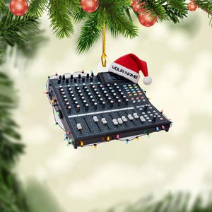 May include: A black audio mixer Christmas ornament with a red Santa hat that says "YOUR NAME". The ornament is decorated with colorful Christmas lights and is hanging from a gold string. Festive holiday decor.