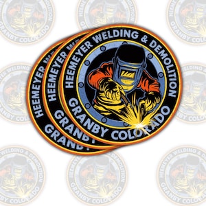 May include: Circular sticker featuring a welder in action, with a welding mask and sparks. The sticker has a black border with the text "HEEMEYER WELDING & DEMOLITION" and "GRANBY COLORADO". The design includes orange, yellow, blue, and black colors.