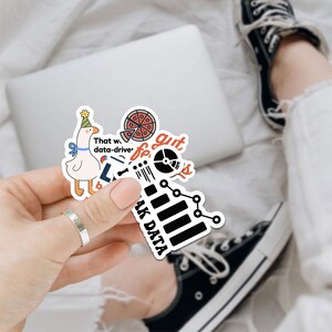 Puede incluir: Un conjunto de pegatinas coloridas con un ganso de dibujos animados, pizza y gr&aacute;ficos relacionados con datos. Las pegatinas tienen textos como "That was data-driven" y "AK DATA". Las pegatinas se sostienen en una mano.