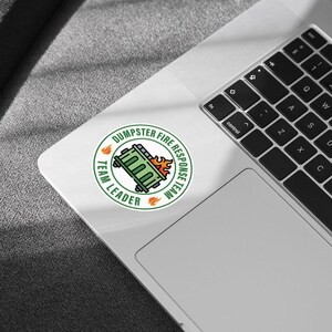 May include: A round sticker with a green and orange design. The sticker features a green dumpster on fire, surrounded by the words "DUMPSTER FIRE RESPONSE TEAM" and "TEAM LEADER" in a circular layout. The sticker is on a laptop.