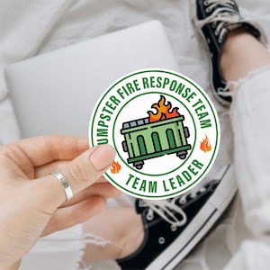May include: A round sticker with the text "DUMPSTER FIRE RESPONSE TEAM" and "TEAM LEADER" encircling a green dumpster on wheels with flames. The sticker has a white background with orange flames.