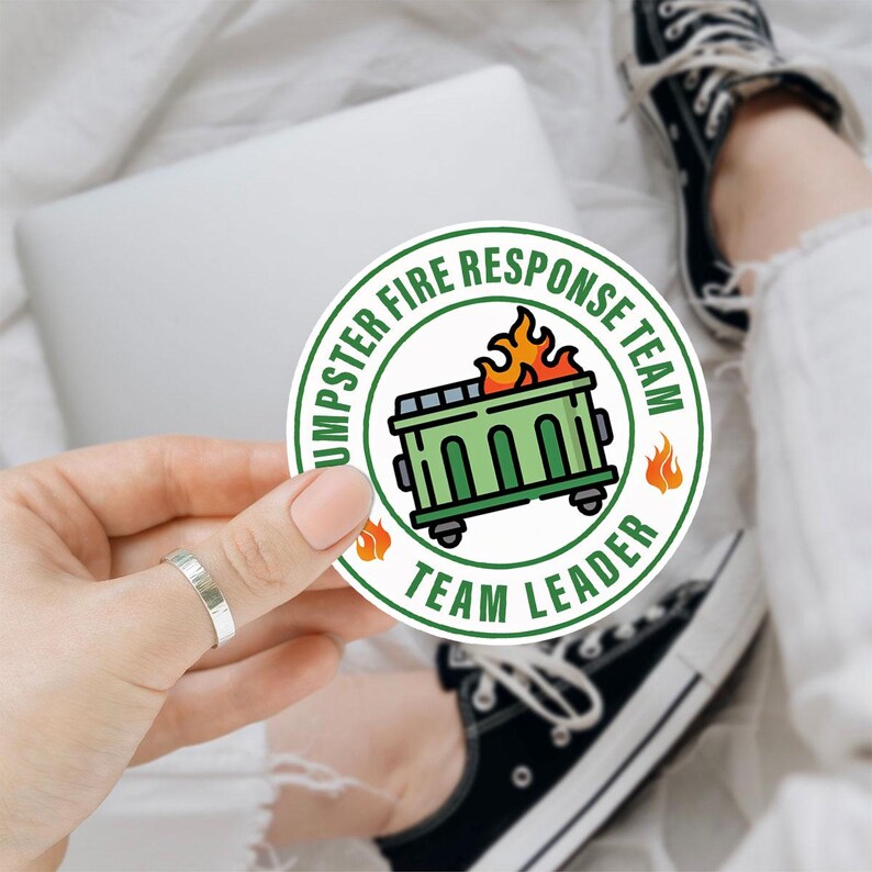 May include: A round sticker with the text "DUMPSTER FIRE RESPONSE TEAM" and "TEAM LEADER" encircling a green dumpster on wheels with flames. The sticker has a white background with orange flames.
