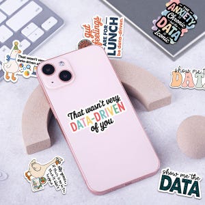Puede incluir: Un tel&eacute;fono inteligente rosa con una pegatina que dice "That wasn't very DATA-DRIVEN of you." Tambi&eacute;n son visibles varias pegatinas con frases relacionadas con datos y un teclado. Las pegatinas tienen varios colores y fuentes.