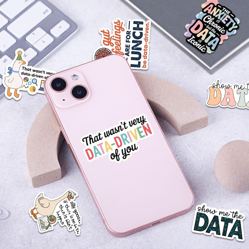 Puede incluir: Un tel&eacute;fono inteligente rosa con una pegatina que dice "That wasn't very DATA-DRIVEN of you." Tambi&eacute;n son visibles varias pegatinas con frases relacionadas con datos y un teclado. Las pegatinas tienen varios colores y fuentes.