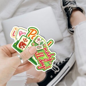 A person's hand holding a sticker with a jig-o-matic and a message, with a pair of sneakers visible in the background.
