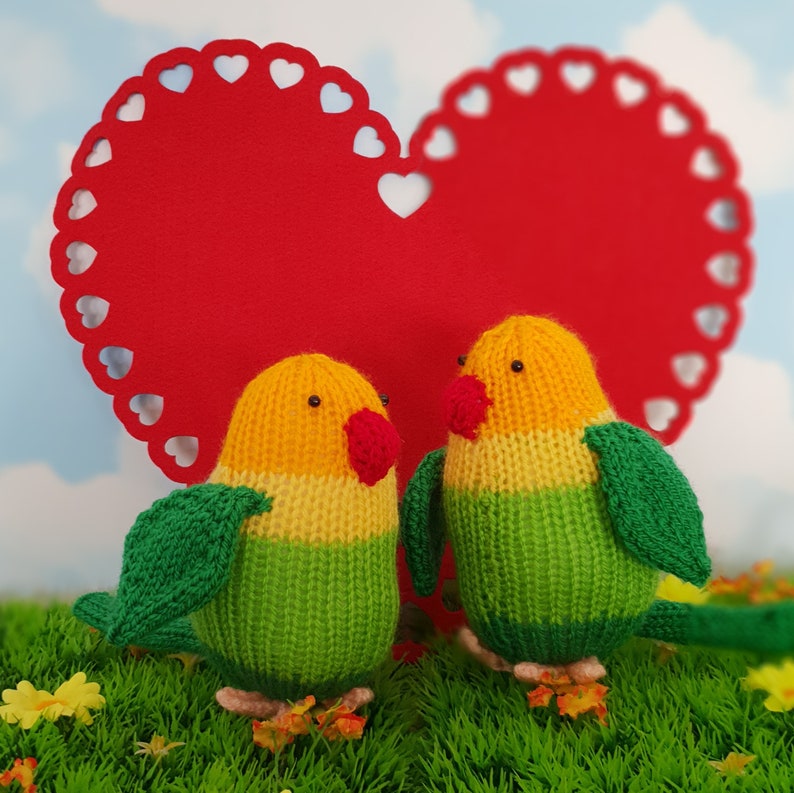 Two colourful knitted lovebirds standing in front of a giant red felt heart.