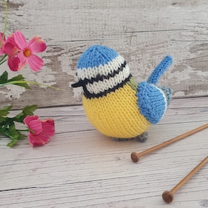 May include: A knitted bluebird toy with a yellow body, blue wings, and a white and black striped head. The bird is sitting on a wooden surface.