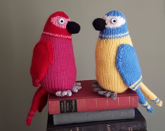 May include: Two knitted bird toys, one red and one yellow and blue, are perched on a stack of books. The red bird has a black beak and the yellow and blue bird has a black beak and white eyes.