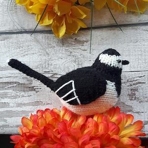 May include: A black and white knitted bird with a long tail, sitting on a bed of orange and yellow flowers.