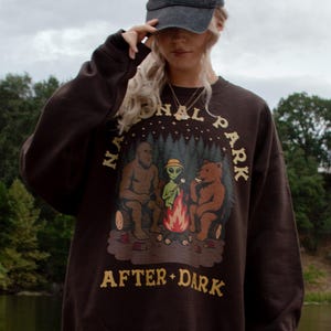 Pode incluir: Uma camisola de gola redonda castanho-escura com um gr&aacute;fico de Bigfoot, um alien&iacute;gena e um urso sentados &agrave; volta de uma fogueira com o texto NATIONAL PARK AFTER DARK, combinada com cal&ccedil;as de ganga azul-claras rasgadas e um bon&eacute; cinzento-escuro.