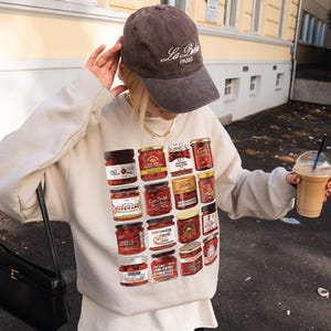 May include: A sand-colored sweatshirt featuring a collage of jarred food products. The person is wearing a brown baseball cap with "La Paris" text and holding a coffee drink. A black purse hangs from the shoulder.
