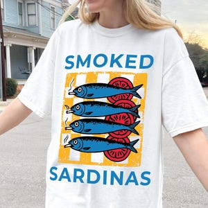 May include: A white t-shirt with a graphic of a stack of smoked sardines on a yellow and white striped background. The text 'SMOKED' is above the sardines and the text 'SARDINAS' is below the sardines.