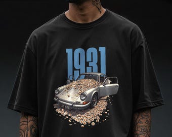 Puede incluir: Camiseta negra con un gráfico de un coche plateado desbordante de flores. El número "1931" está impreso en azul encima del coche. La camiseta tiene mangas cortas y un corte holgado.