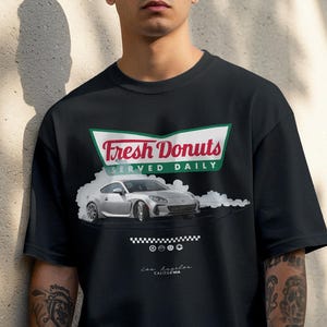 Op de afbeelding: Zwart T-shirt met een afbeelding van een zilveren sportwagen die drift, met de tekst "Fresh Donuts Served Daily" in een groen en wit bord. Onder de auto staan vier kleine cirkelvormige ontwerpen en de tekst "Los Angeles, California".