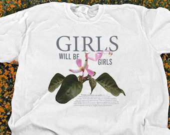 May include: White t-shirt featuring the text "GIRLS WILL BE GIRLS" in grey. A pink and green floral design is positioned below the text. The shirt is displayed against a backdrop of orange and green flowers.