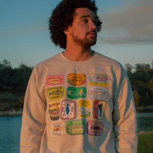 May include: A beige crewneck sweatshirt featuring a grid of colorful rectangular sardine tin labels. The graphic print includes various vintage-style illustrations of fish, text, and brand logos in shades of red, blue, green, and yellow on the front.